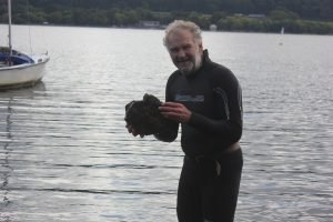 Ian Hughes in a wetsuit standing in a lake, holding a rock, smiling to camera.