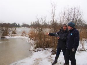 Two women standing by an icy pond, one pointing at the water